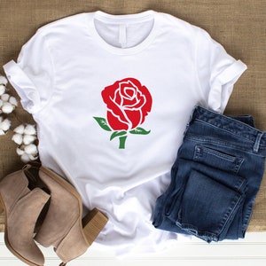 K&ouml;nnte beinhalten: Wei&szlig;es T-Shirt mit roter Rosen-Grafik, gr&uuml;nen Bl&auml;ttern und Stiel. Kombiniert mit blauen Jeans und beigen Stiefeletten, ergibt sich ein l&auml;ssiger Alltagslook. Das Bild zeigt Mode und Bekleidung.