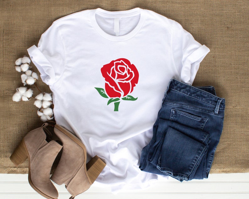 K&ouml;nnte beinhalten: Wei&szlig;es T-Shirt mit roter Rosen-Grafik, gr&uuml;nen Bl&auml;ttern und Stiel. Kombiniert mit blauen Jeans und beigen Stiefeletten, ergibt sich ein l&auml;ssiger Alltagslook. Das Bild zeigt Mode und Bekleidung.