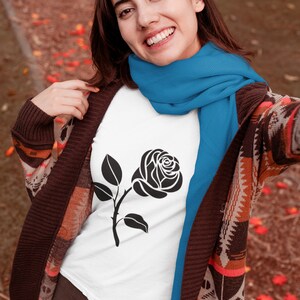 May include: A white t-shirt featuring a black rose graphic. The person is wearing a blue scarf and a brown patterned cardigan. The background has a blurred autumn setting with fallen leaves.