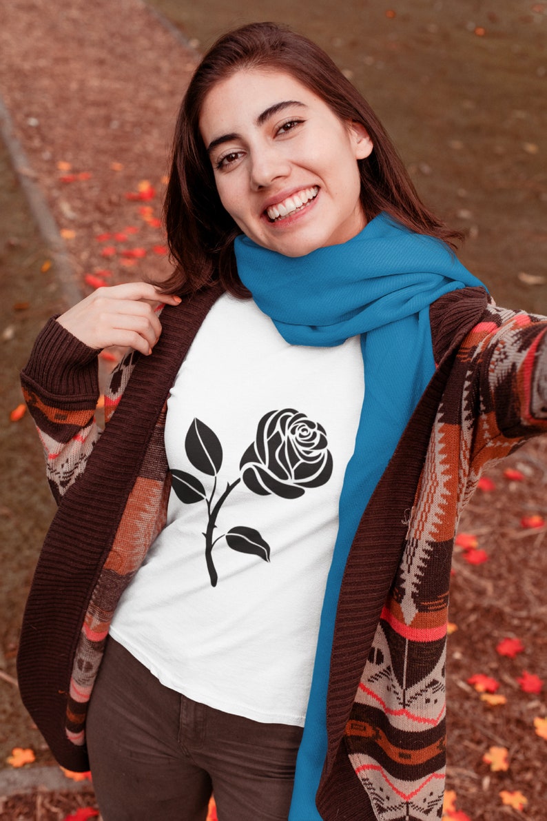 May include: A white t-shirt featuring a black rose graphic. The person is wearing a blue scarf and a brown patterned cardigan. The background has a blurred autumn setting with fallen leaves.