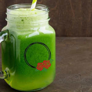 K&ouml;nnte beinhalten: Ein Einmachglas gef&uuml;llt mit einem leuchtend gr&uuml;nen Smoothie, garniert mit Schaum und einem gestreiften Strohhalm. Ein schwarzes kreisf&ouml;rmiges Design mit roten Rosen verziert das Glas. Der Hintergrund ist dunkelbraun.