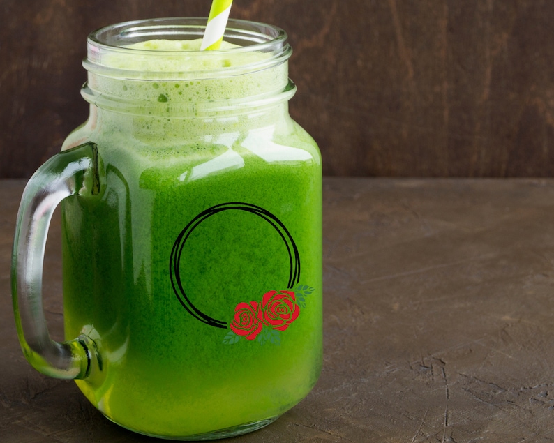 K&ouml;nnte beinhalten: Ein Einmachglas gef&uuml;llt mit einem leuchtend gr&uuml;nen Smoothie, garniert mit Schaum und einem gestreiften Strohhalm. Ein schwarzes kreisf&ouml;rmiges Design mit roten Rosen verziert das Glas. Der Hintergrund ist dunkelbraun.