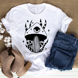 May include: White t-shirt with a black and white illustration of a mushroom with an eye. The mushroom has a swirling pattern and stars around it.
