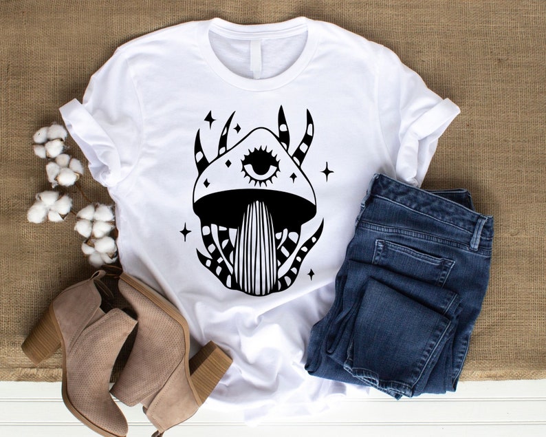 May include: White t-shirt with a black and white illustration of a mushroom with an eye. The mushroom has a swirling pattern and stars around it.