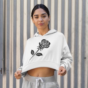 May include: White cropped hoodie featuring a black rose graphic on the front. The hoodie has a drawstring hood and long sleeves. The model is wearing gray sweatpants. The background is a corrugated metal wall.
