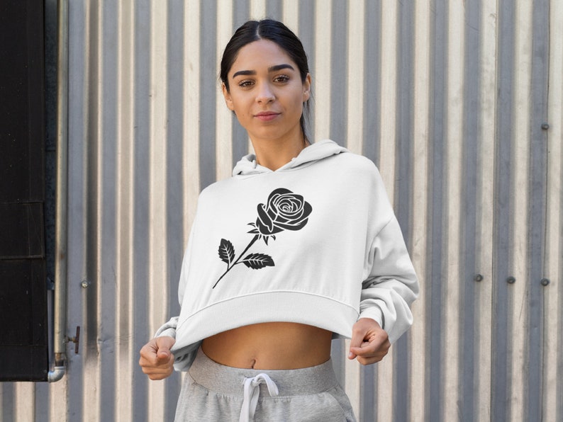 May include: White cropped hoodie featuring a black rose graphic on the front. The hoodie has a drawstring hood and long sleeves. The model is wearing gray sweatpants. The background is a corrugated metal wall.