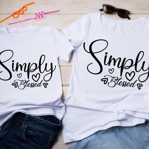 Puede incluir: Dos camisetas blancas con el texto "Simply Blessed" en letras cursivas negras y pequeños corazones. Las camisetas están colocadas sobre una superficie de madera.