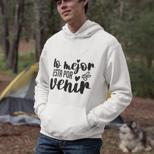 Op de afbeelding: Witte hoodie met de tekst "lo mejor esta por venir" in zwart schrift. De hoodie heeft een voorzak en een capuchon met trekkoord. De persoon op de foto draagt een bruine pet en een blauwe jeans.