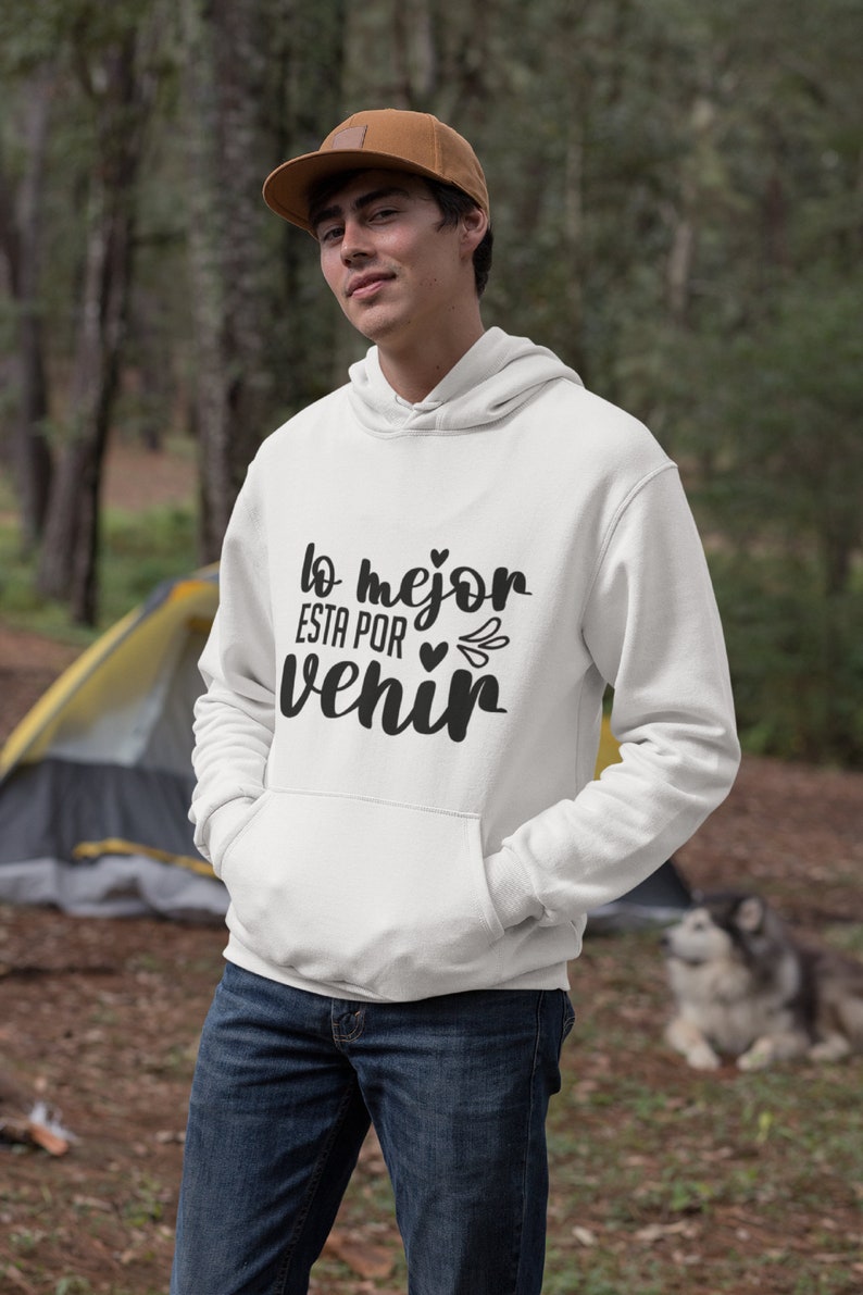 Op de afbeelding: Witte hoodie met de tekst "lo mejor esta por venir" in zwart schrift. De hoodie heeft een voorzak en een capuchon met trekkoord. De persoon op de foto draagt een bruine pet en een blauwe jeans.