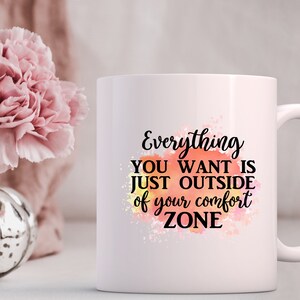 K&ouml;nnte beinhalten: Wei&szlig;e Keramiktasse mit einem motivierenden Zitat: "Alles, was du willst, ist direkt au&szlig;erhalb deiner Komfortzone." Das Zitat ist in schwarzer Schrift auf einem Aquarellhintergrund in Orange, Gelb und Rosa geschrieben.