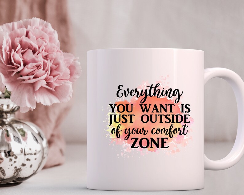 K&ouml;nnte beinhalten: Wei&szlig;e Keramiktasse mit einem motivierenden Zitat: "Alles, was du willst, ist direkt au&szlig;erhalb deiner Komfortzone." Das Zitat ist in schwarzer Schrift auf einem Aquarellhintergrund in Orange, Gelb und Rosa geschrieben.