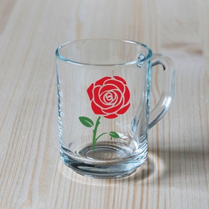 K&ouml;nnte beinhalten: Ein klarer Glasbecher mit Henkel, verziert mit einem roten Rosenmuster mit gr&uuml;nen Bl&auml;ttern und Stiel. Der Becher ist transparent, so dass der Hintergrund sichtbar ist.