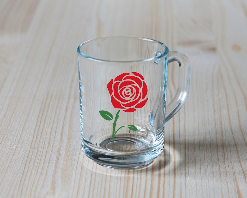 K&ouml;nnte beinhalten: Ein klarer Glasbecher mit Henkel, verziert mit einem roten Rosenmuster mit gr&uuml;nen Bl&auml;ttern und Stiel. Der Becher ist transparent, so dass der Hintergrund sichtbar ist.