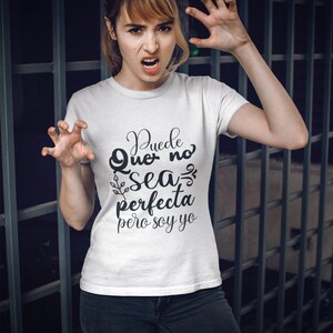 Op de afbeelding: Wit T-shirt met de Spaanse tekst "Puede Que no sea perfecta pero soy yo" in zwart script. De persoon draagt een zwarte jeans en maakt een klauwende beweging met haar handen.