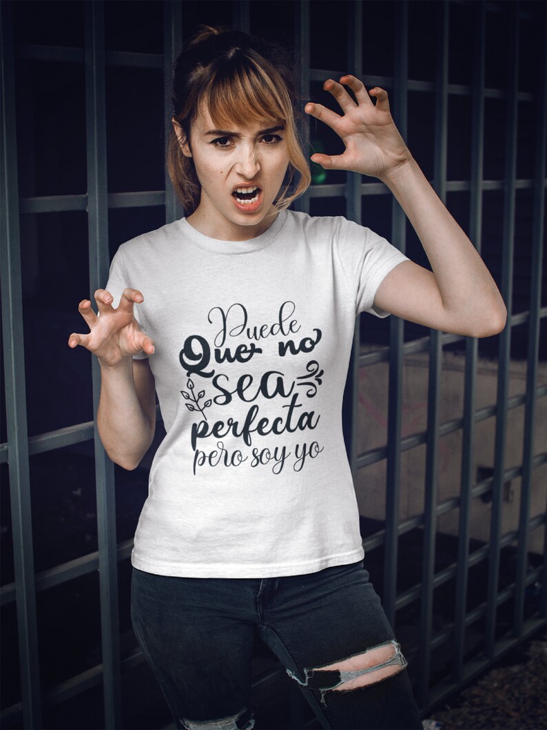 Op de afbeelding: Wit T-shirt met de Spaanse tekst "Puede Que no sea perfecta pero soy yo" in zwart script. De persoon draagt een zwarte jeans en maakt een klauwende beweging met haar handen.