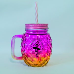 May include: A pink and orange glass mason jar with a handle and a straw. The jar has a diamond pattern and a black and white graphic of a mushroom with an eye on top.