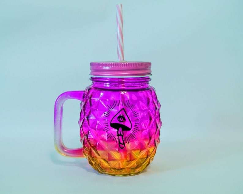 May include: A pink and orange glass mason jar with a handle and a straw. The jar has a diamond pattern and a black and white graphic of a mushroom with an eye on top.