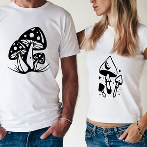 May include: Two white t-shirts with black mushroom illustrations. One shirt features a cluster of three mushrooms with a crescent moon on the top of one. The other shirt features a cluster of three mushrooms with white dots on the top of one.