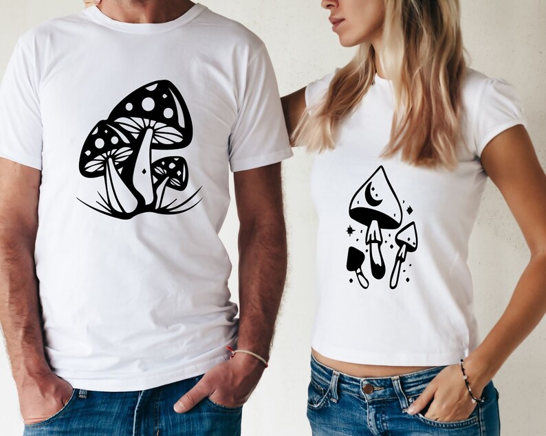 May include: Two white t-shirts with black mushroom illustrations. One shirt features a cluster of three mushrooms with a crescent moon on the top of one. The other shirt features a cluster of three mushrooms with white dots on the top of one.