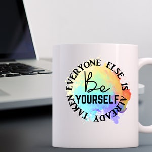 K&ouml;nnte beinhalten: Wei&szlig;e Keramik-Kaffeetasse mit einem farbenfrohen Aquarelldesign und dem Text "Be Yourself Everyone Else Is Already Taken".