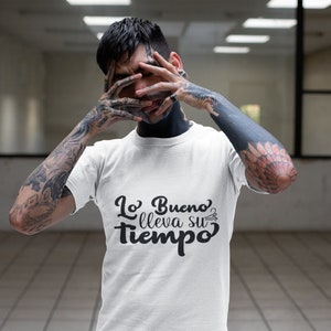 Op de afbeelding: Wit T-shirt met de Spaanse zin "Lo Bueno lleva su tiempo" in zwart schrift. Het shirt wordt gedragen door een persoon met tatoeages op armen en nek.