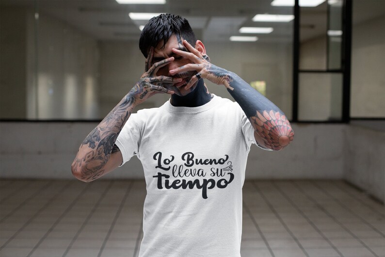 Op de afbeelding: Wit T-shirt met de Spaanse zin "Lo Bueno lleva su tiempo" in zwart schrift. Het shirt wordt gedragen door een persoon met tatoeages op armen en nek.