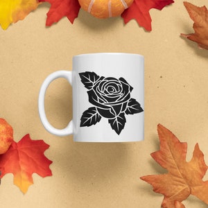 May include: White ceramic mug featuring a black rose design. The mug has a curved handle and a smooth surface. The mug is surrounded by fall-themed decorations, including leaves, pumpkins, and pinecones.