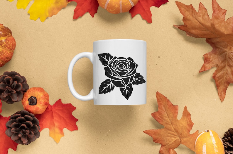 May include: White ceramic mug featuring a black rose design. The mug has a curved handle and a smooth surface. The mug is surrounded by fall-themed decorations, including leaves, pumpkins, and pinecones.