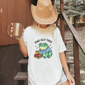 Może przedstawiać: Biały t-shirt z krótkim rękawem z grafiką przedstawiającą zieloną żabę w okularach i niebieskiej koszuli, siedzącą obok stosu książek z jabłkiem na wierzchu. Tekst nad żabą brzmi TOAD-ALLY TIRED i jest napisany czarnymi wielkimi literami.