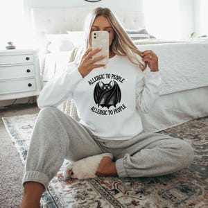May include: A white long-sleeve sweatshirt featuring a black graphic of a bat with wings wrapped around its body. The text "ALLERGIC TO PEOPLE" is printed in a curved black font above and below the bat illustration on the centre of the chest.