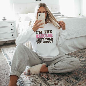 以下が含まれることがあります： 茶色とピンクの文字で「I'M THE REGULAR THEY TOLD YOU ABOUT」とプリントされた白い長袖のクルーネックトレーナー。グレーのスウェットパンツと白いふわふわしたスリッパを合わせています。