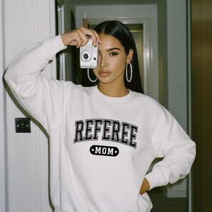 Könnte beinhalten: Ein weißes langärmeliges Sweatshirt mit Rundhalsausschnitt und dem schwarzen Schriftzug REFEREE in einer College-Schriftart, der über einer schwarzen, pillenförmigen Grafik mit dem Wort MOM und zwei kleinen weißen Sternen gewölbt ist.
