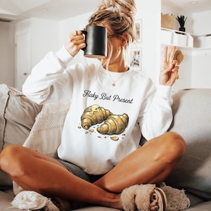 May include: A white long-sleeve sweatshirt featuring a graphic of two golden-brown croissants with a pat of butter and the text Flaky But Present printed in black script. The garment has a crew neckline and ribbed cuffs, showcasing a casual style.
