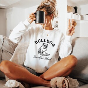 Può includere: Una felpa bianca a maniche lunghe con una grafica nera di un bulldog con gli occhiali, seduto con una tazza di caffè su un giornale. Il testo sopra il cane recita BULLDOG e il testo sottostante Breakfast Blend in un carattere corsivo nero.