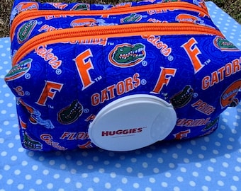 May include: A rectangular fabric pouch featuring a blue background with an orange and green Florida Gators sports team print. The pouch has two orange zippers and a white plastic Huggies brand flip-top lid centered on the front panel.