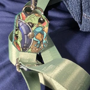 May include: A light green strap with a silver clasp and a small, colorful charm featuring a cartoon character. The charm is attached to a denim fabric piece with silver rivets. The background is a dark blue fabric.