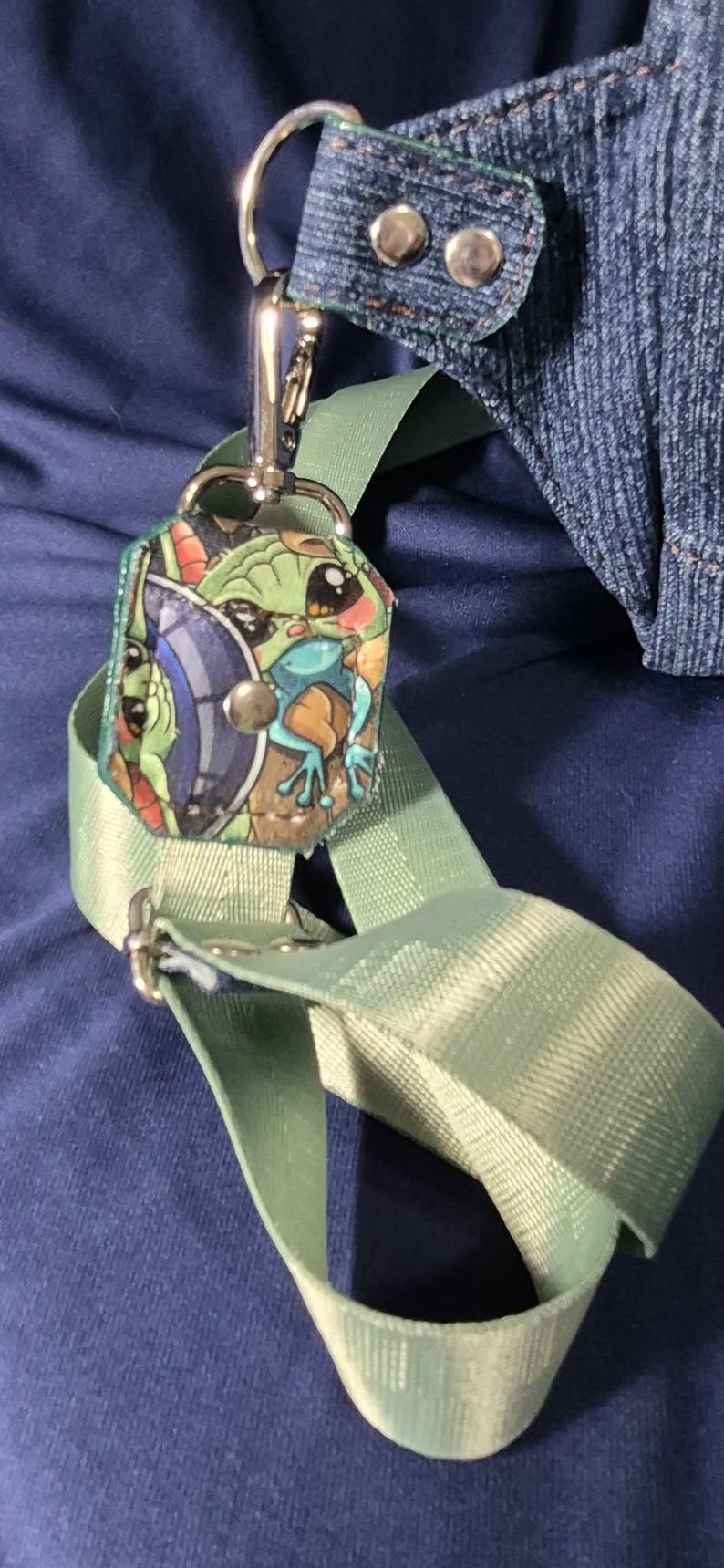 May include: A light green strap with a silver clasp and a small, colorful charm featuring a cartoon character. The charm is attached to a denim fabric piece with silver rivets. The background is a dark blue fabric.
