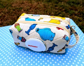 May include: A quilted, cream-colored fabric pouch featuring a repeating pattern of Sesame Street characters. The rectangular case has two zippered compartments, a side carrying strap, and a white plastic HUGGIES branded flip-top dispenser.