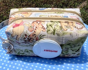 May include: A rectangular quilted fabric pouch featuring a Winnie the Pooh and Tigger print in soft pastel colors. The pouch has two beige zippered compartments and a white plastic Huggies branded flip-top dispenser lid on the front side.