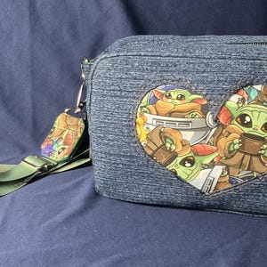 May include: Denim crossbody bag with a heart-shaped graphic featuring a cartoon character. The bag has a green strap and a small charm. The bag is approximately 8 inches wide and 6 inches tall.