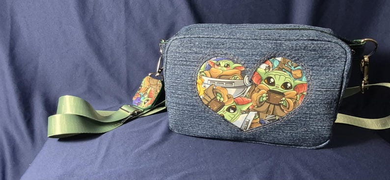 May include: Denim crossbody bag with a heart-shaped graphic featuring a cartoon character. The bag has a green strap and a small charm. The bag is approximately 8 inches wide and 6 inches tall.