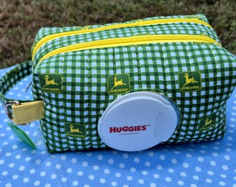 May include: A quilted fabric pouch featuring a green and white gingham pattern with the John Deere logo and a white plastic Huggies wipe dispenser lid on the front. The pouch includes a yellow zipper closure and a yellow side strap with a metal ring.