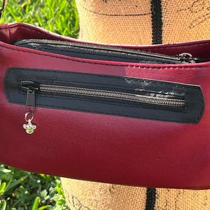 Puede incluir: Un peque&ntilde;o bolso bandolera de color burdeos con una cremallera negra y un abalorio de abeja plateado. El bolso tiene cierre de solapa y una correa de cadena.