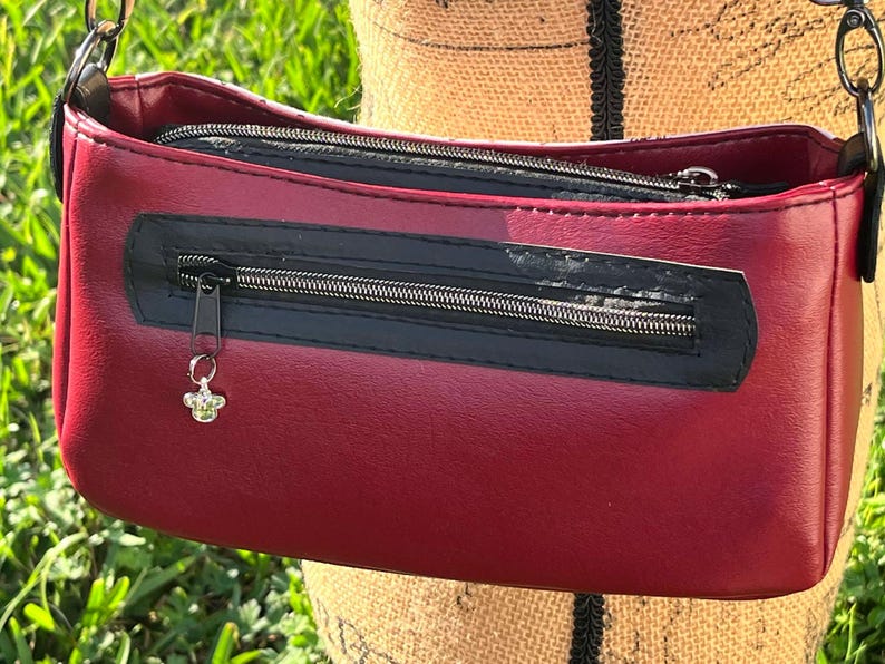 Puede incluir: Un peque&ntilde;o bolso bandolera de color burdeos con una cremallera negra y un abalorio de abeja plateado. El bolso tiene cierre de solapa y una correa de cadena.