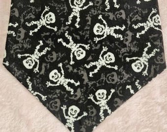 May include: A black and white Halloween-themed pet bandana. The bandana features a repeating pattern of white skeleton figures and gray skulls on a black background. The bandana is triangular in shape.