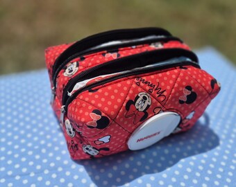 May include: A red quilted fabric pouch featuring a repeating Minnie Mouse pattern with white polka dots. The rectangular bag has two black zippered compartments and a white plastic flip-top opening on the front with the word Huggies printed on it.