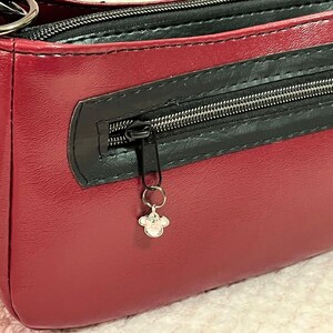 Puede incluir: Un bolso de mano de cuero burdeos con una cremallera negra y un charm plateado. El bolso tiene un asa superior y una cremallera.