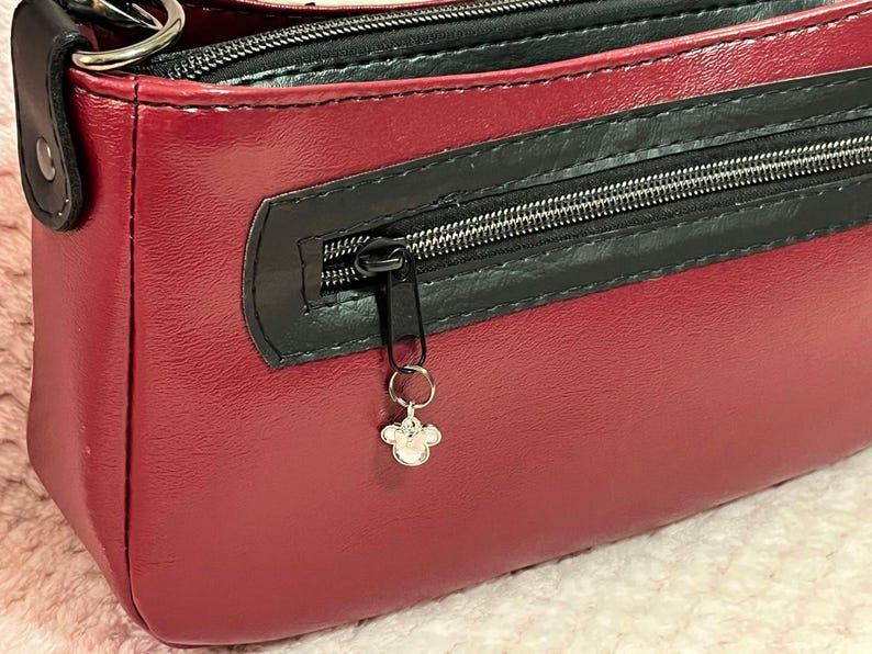 Puede incluir: Un bolso de mano de cuero burdeos con una cremallera negra y un charm plateado. El bolso tiene un asa superior y una cremallera.