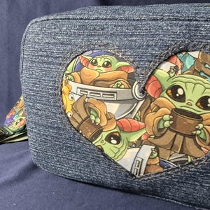 May include: Denim-style crossbody bag featuring a heart-shaped patch with Grogu illustrations. The bag has a green strap and a colorful patterned accent. The Grogu design includes the character holding a cup and in a pod.