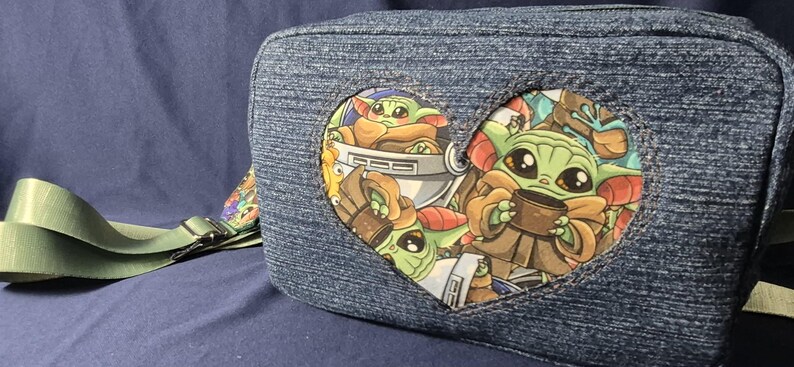 May include: Denim-style crossbody bag featuring a heart-shaped patch with Grogu illustrations. The bag has a green strap and a colorful patterned accent. The Grogu design includes the character holding a cup and in a pod.
